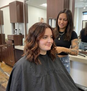 A smiling client with medium-length brunette hair styled in soft, beachy waves sitting in a salon chair at Garbo's Salon and Spa.
