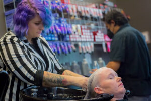A stylist with purple hair and tattoos washes a guest's hair at a professional salon shampoo bowl station, preparing their hair for a premium service at Garbo's Salon.